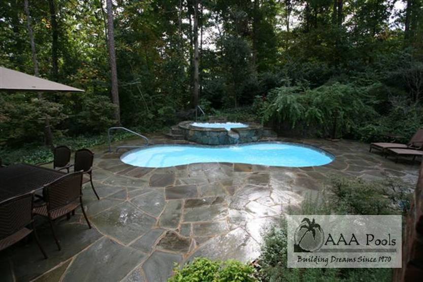 Fiberglass pool in a forested backyard with stone patio and hot tub