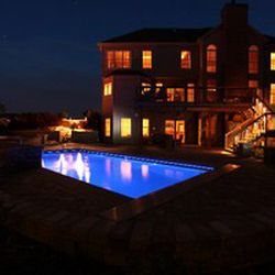 Nighttime view of a rectangular fiberglass pool with dark blue water and warm house glow